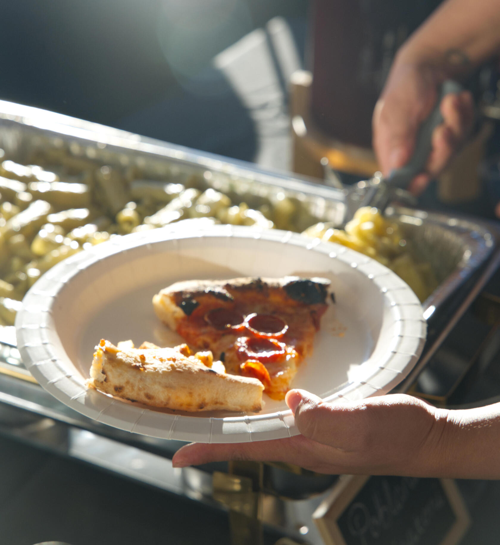 some one serving themselves pasta holding a plate with two slices of pizza